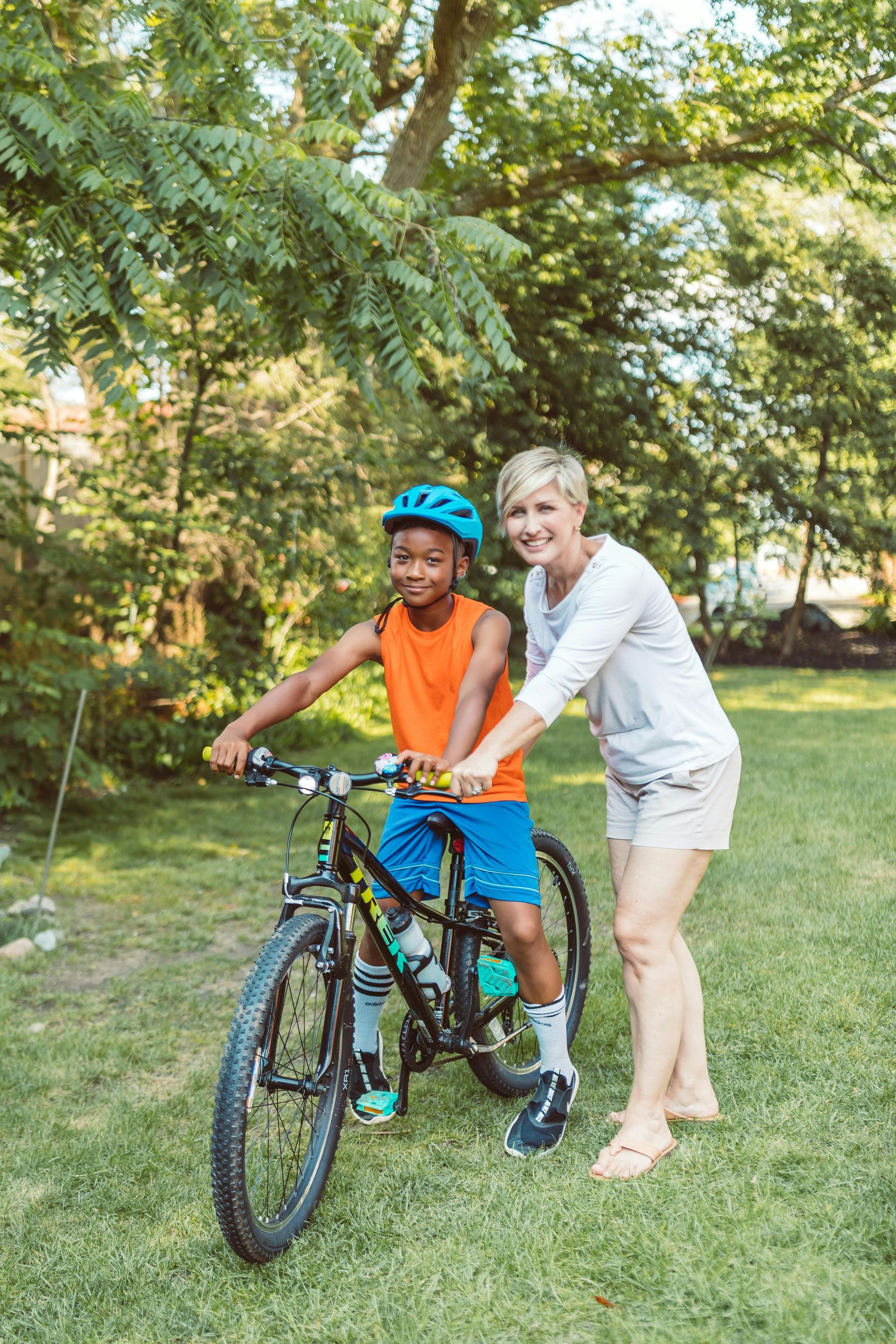 Woman helps a young boy learn to ride a bike in a grassy park.