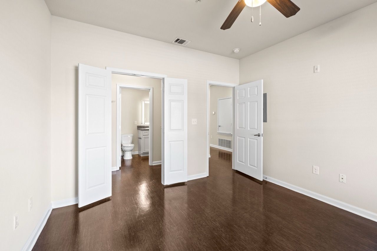 Empty bedroom with white doors open to a bathroom and an adjoining room; dark wood floors and a ceiling fan.