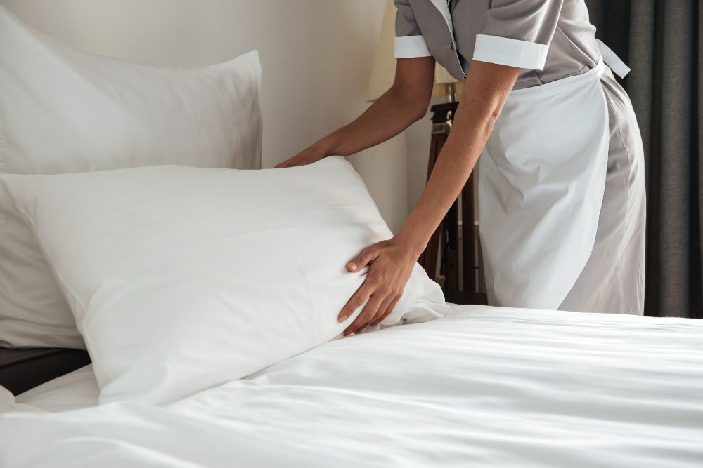 Uma funcionária da limpeza está arrumando uma cama com lençóis e travesseiros brancos.pousada-dos-guias