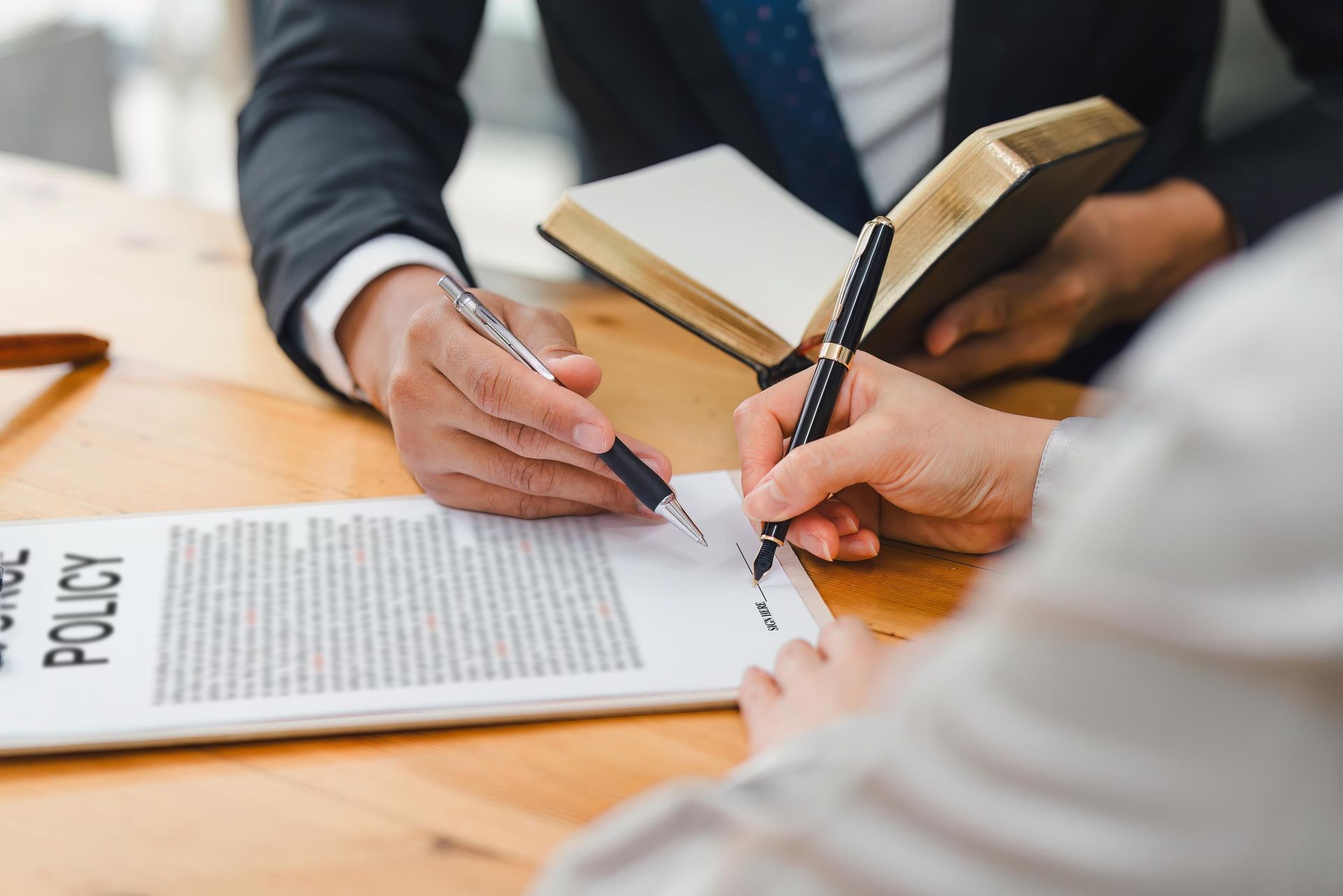 Person signing a document, guided by another person's pen; a book is open nearby.