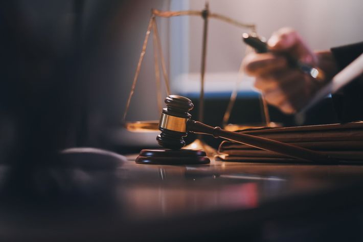 Gavel on wooden table with scales, symbolizing law and justice. A person's hand is visible in the background.