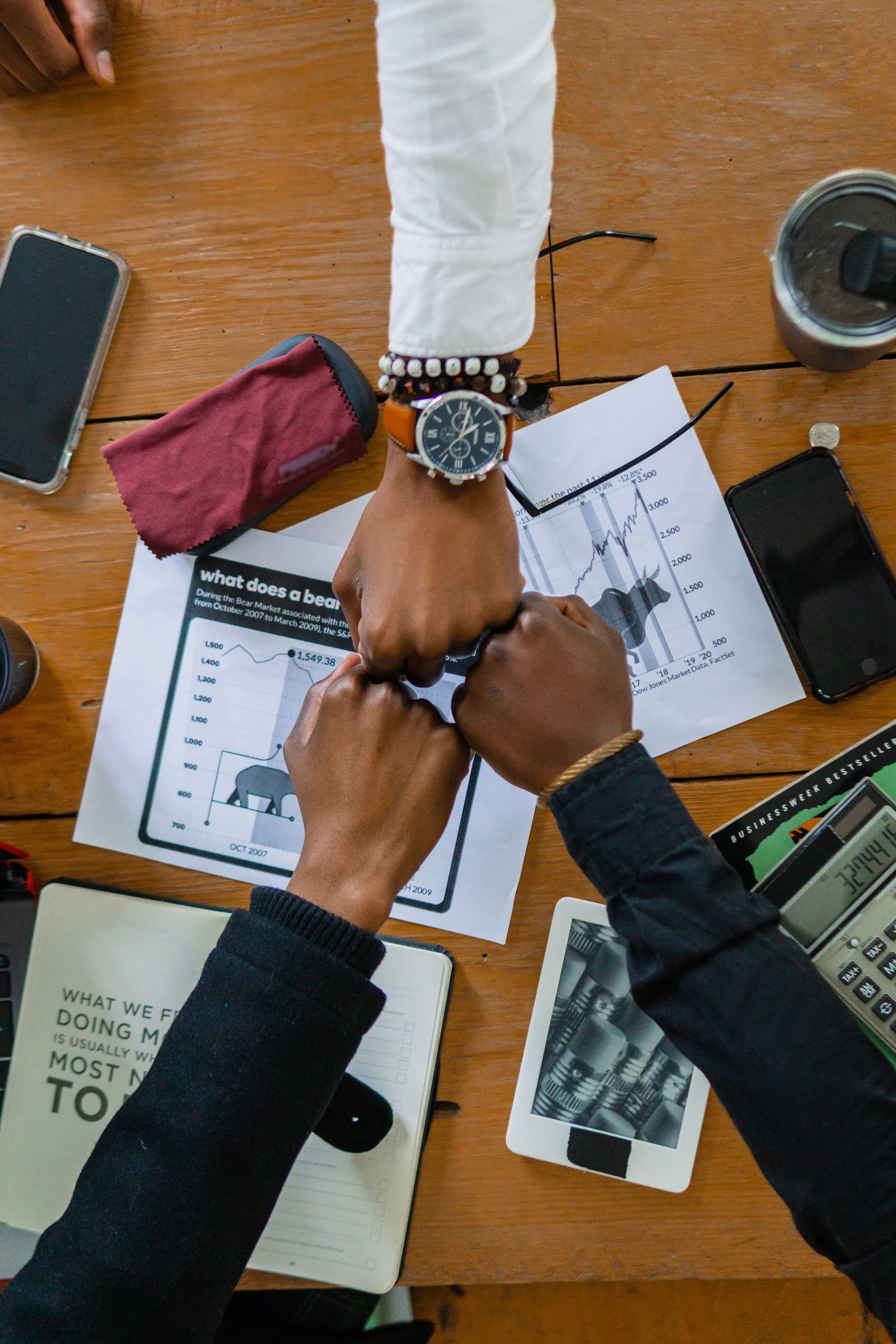 Two people stack hands over papers at a wooden table with notebooks and a phone nearby.