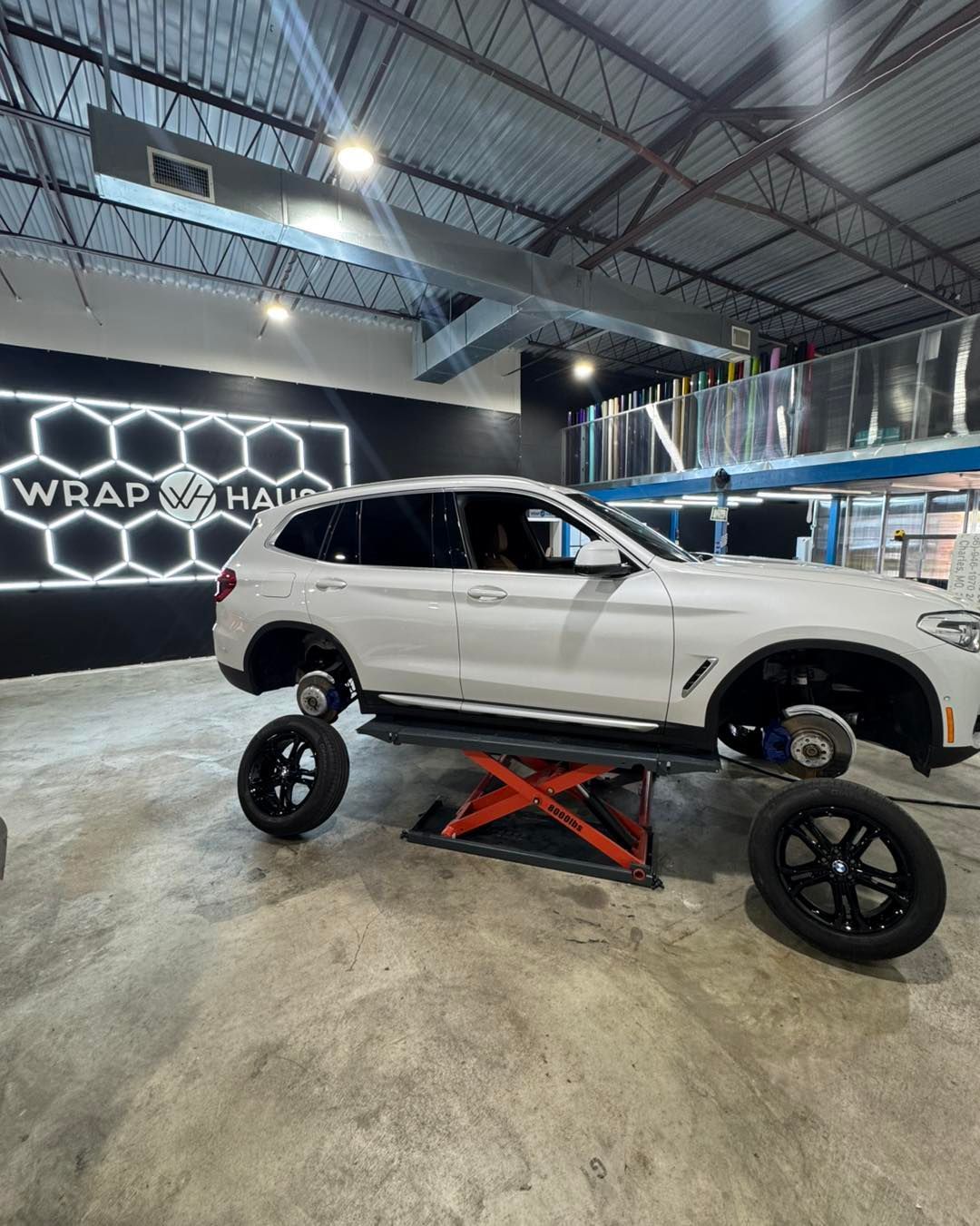 White SUV on a lift in an auto shop, with large black wheels nearby and a Wrap Mafia sign in the background