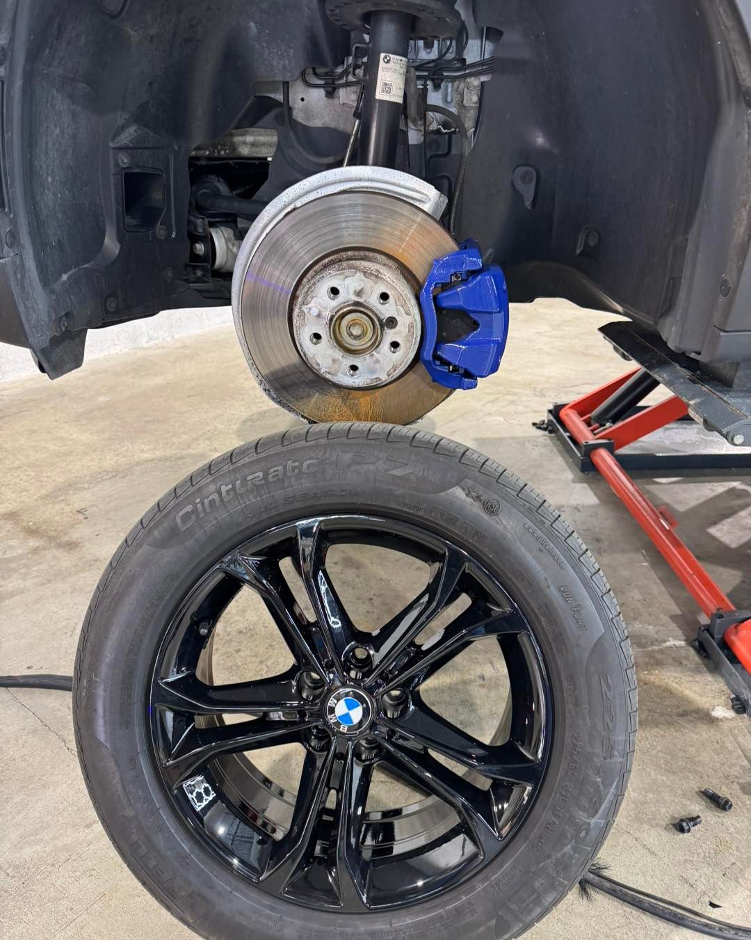Car on a jack with front wheel removed, showing brake rotor and blue caliper beside a black wheel.