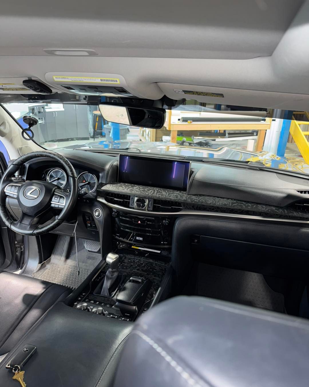 Car interior with black leather seats, steering wheel, dashboard, and center console; garage equipment visible through windshield