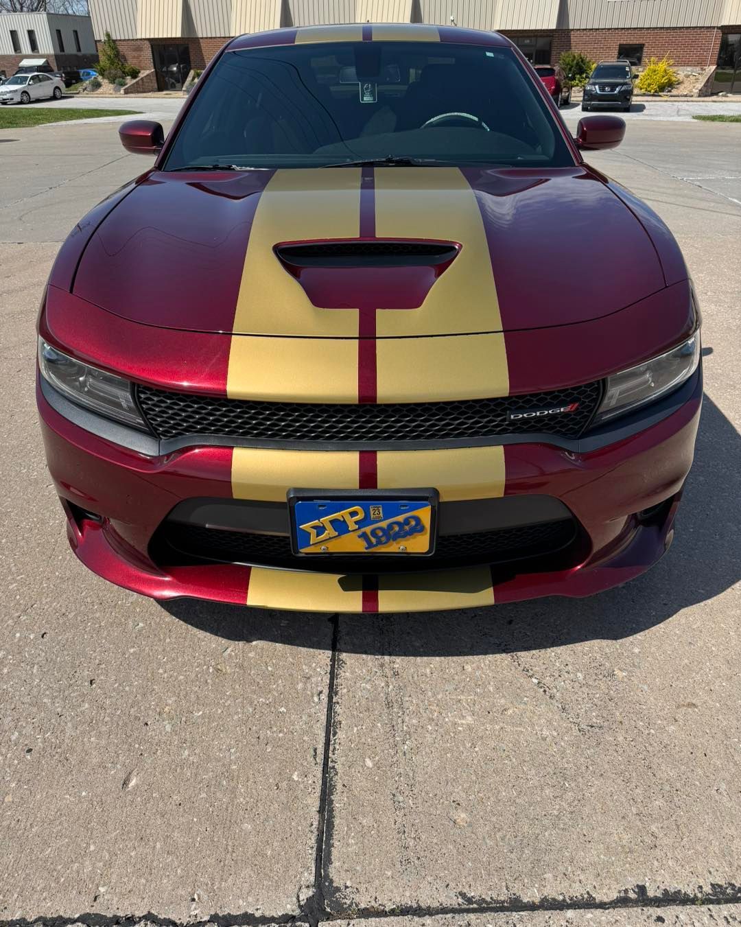 Maroon Dodge Charger with gold racing stripe parked on pavement in front of buildings