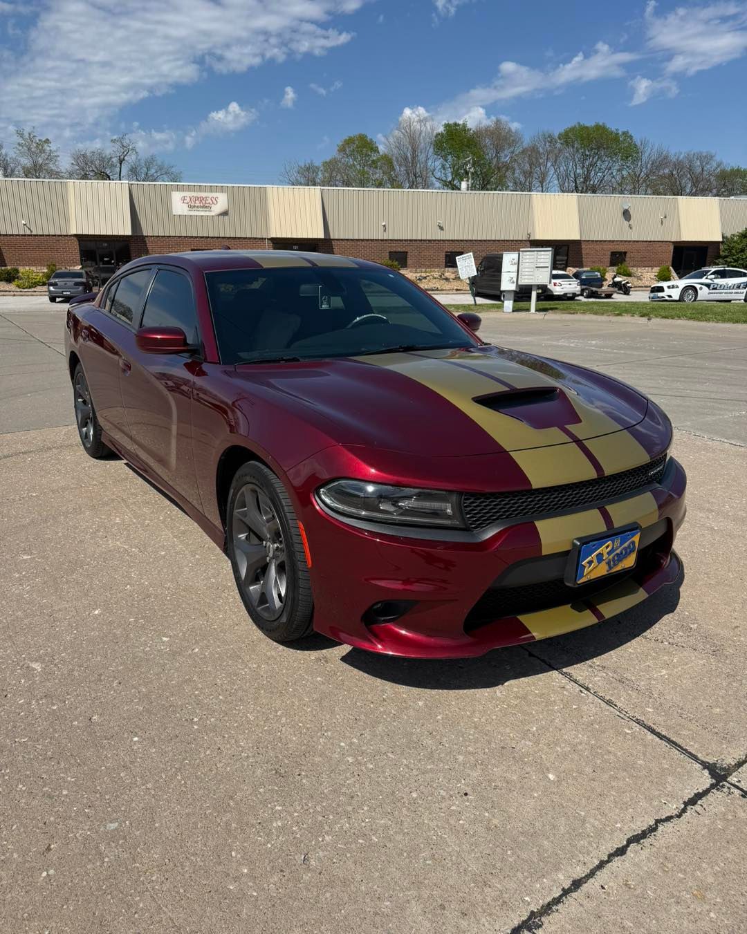 Maroon Dodge Charger with black racing stripes parked in a lot near a tan building under a blue sky