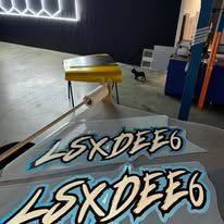 Two LSXDE6 logo stickers on a gray surface in a dim indoor room with a yellow table in the background
