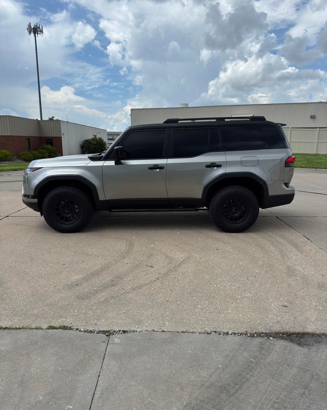 Silver SUV with black roof and wheels parked on a concrete lot under a partly cloudy sky.