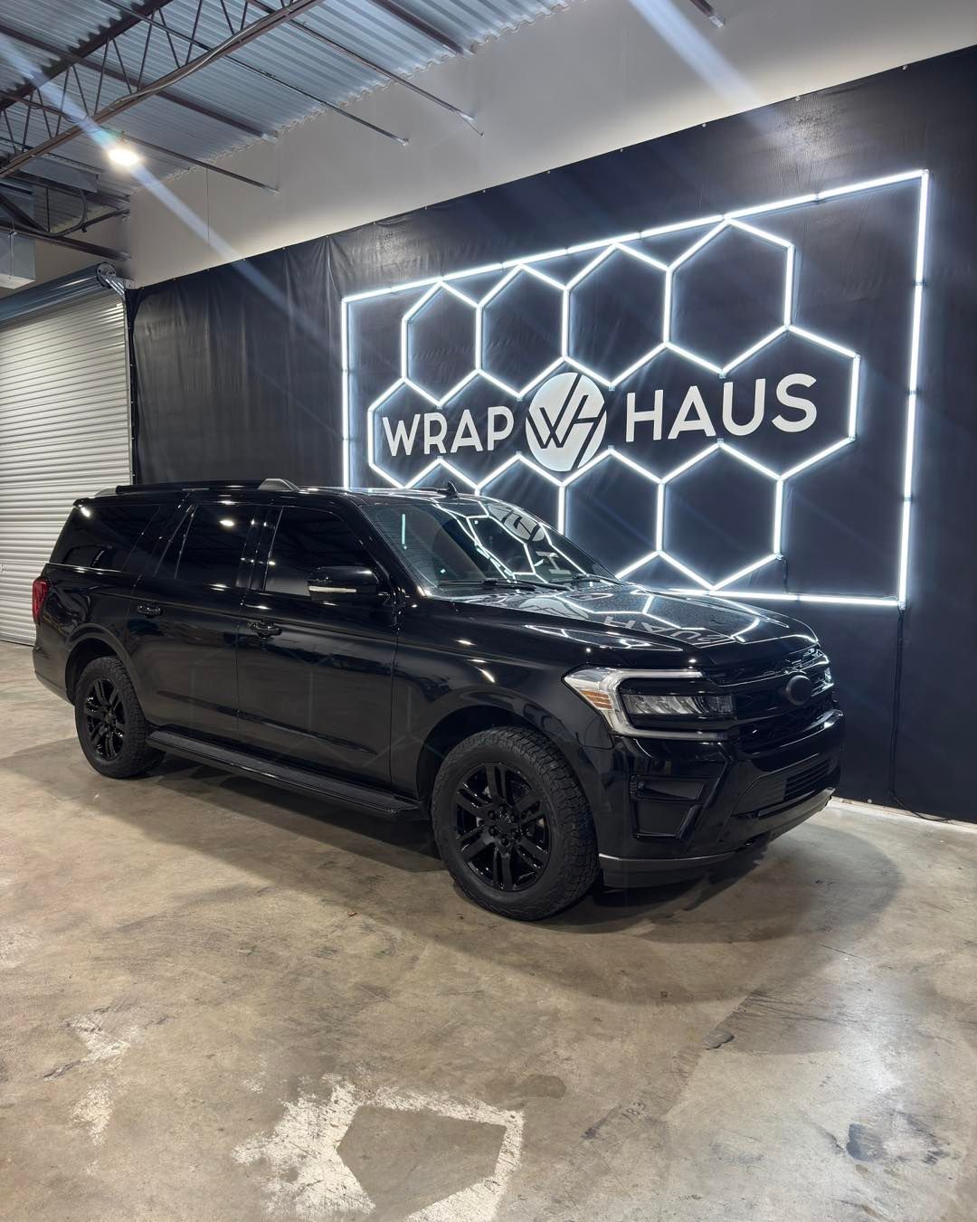 Black SUV inside a wrap shop with a lit “Wrap Haus” sign and hexagon wall design