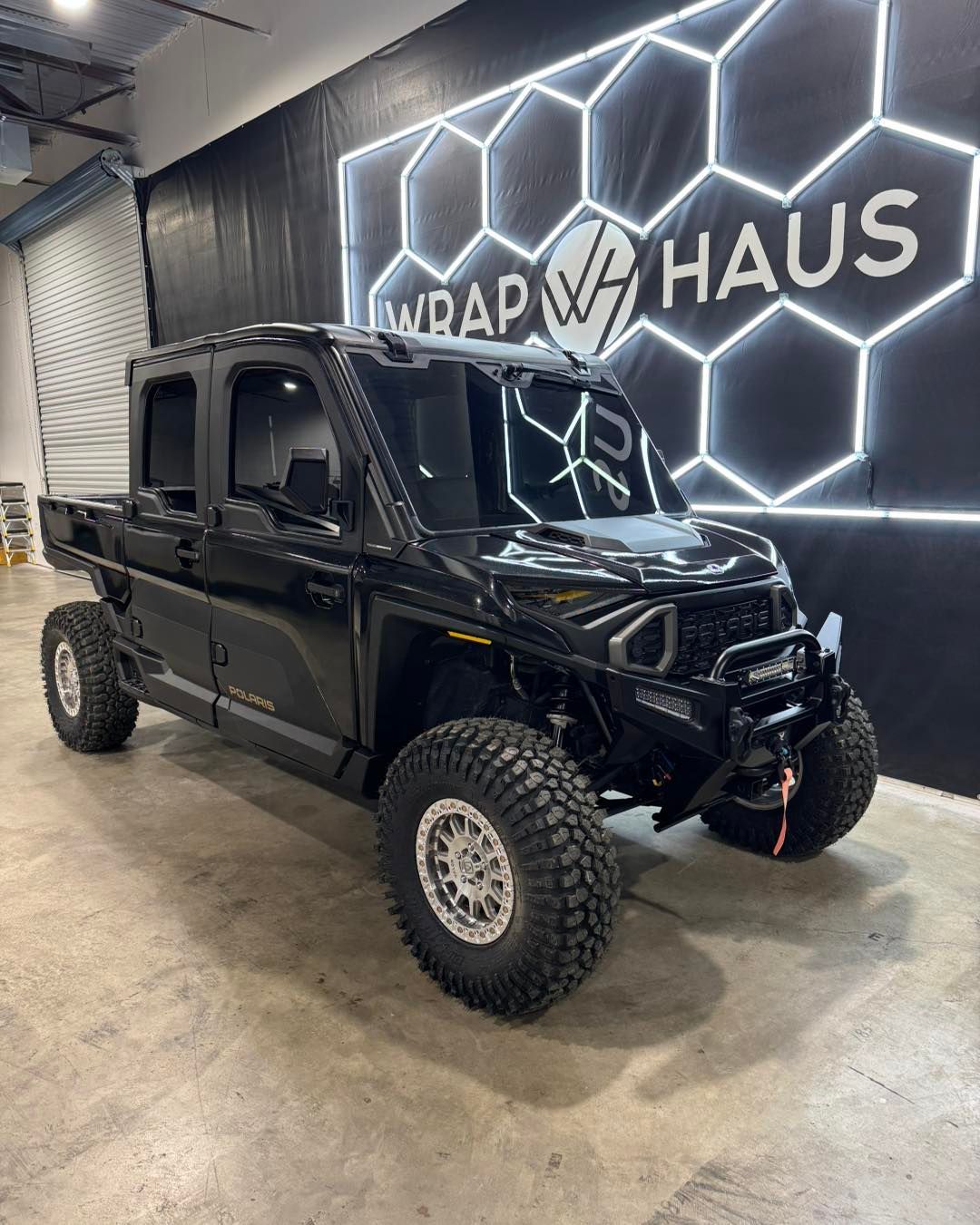 Black lifted pickup truck in a showroom with glowing hexagon wall lights and a “WRAPY HAUS” sign.
