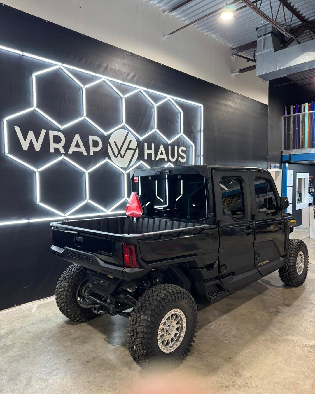 Black lifted Jeep indoors beside a neon honeycomb Wrap Haus wall sign