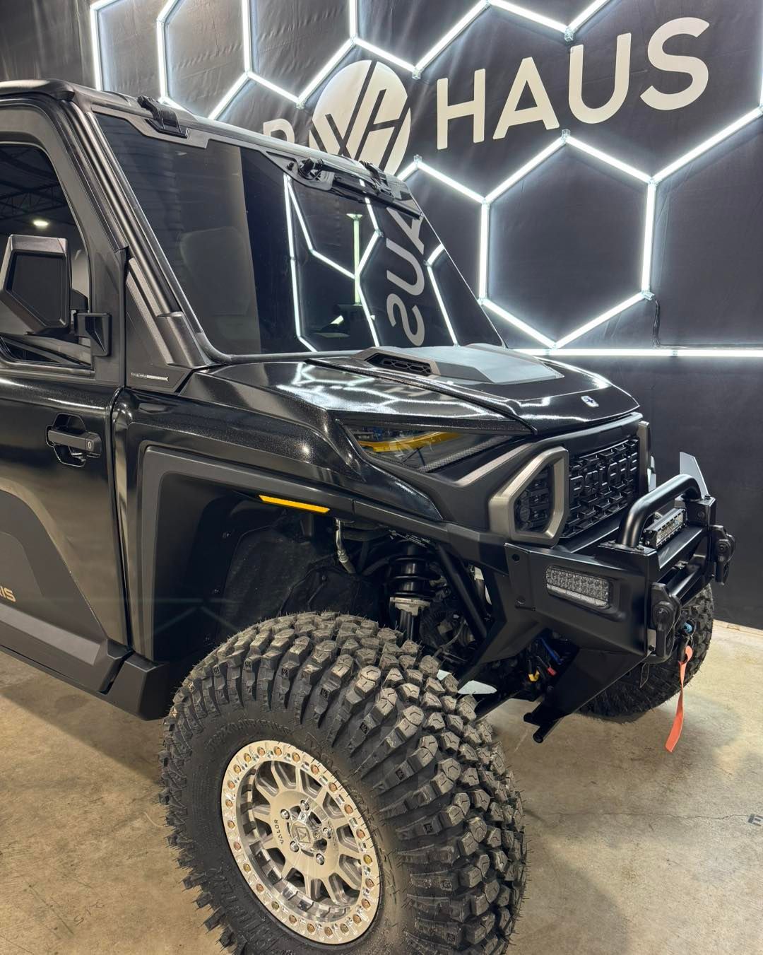 Black off-road truck with large tires displayed indoors against a hexagon-lit wall.