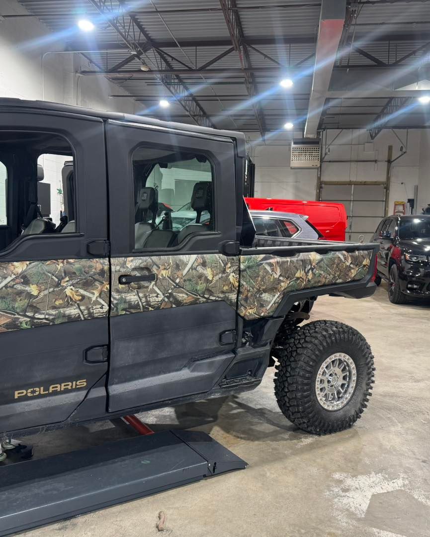 Camouflage Polaris off-road vehicle indoors at an auto show, with a lifted suspension and black step rail