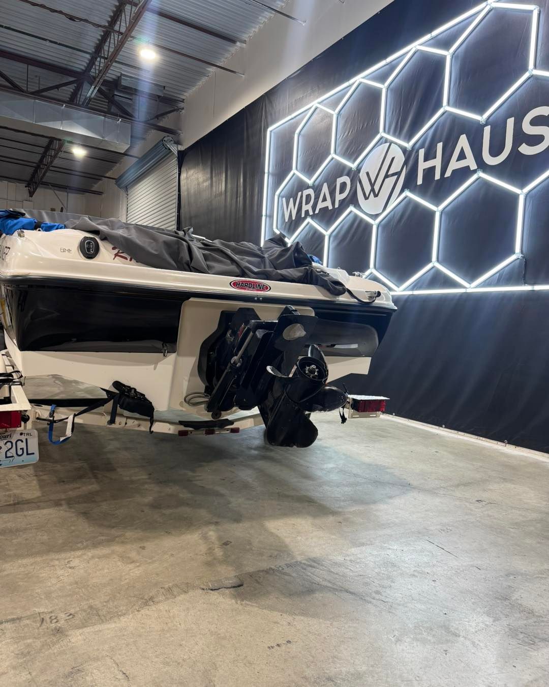White speedboat indoors beside a neon hexagon “WRAP MY HAUS” wall sign