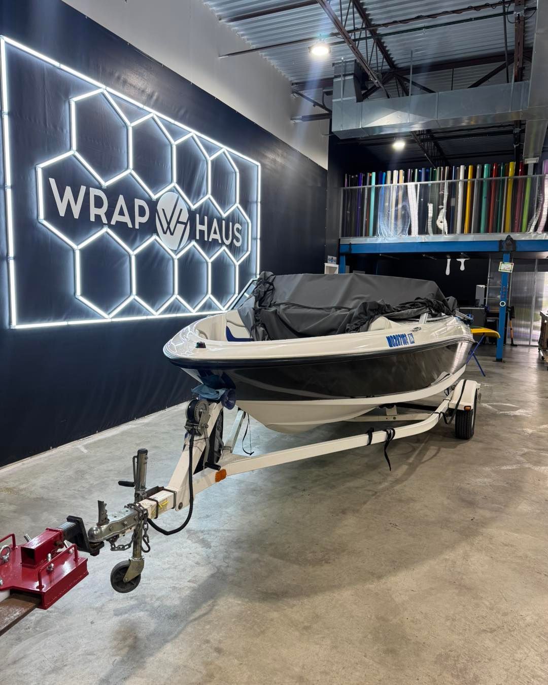 Boat in a workshop under a neon WRAP HOUSE sign, with a tow hitch attached to the trailer.