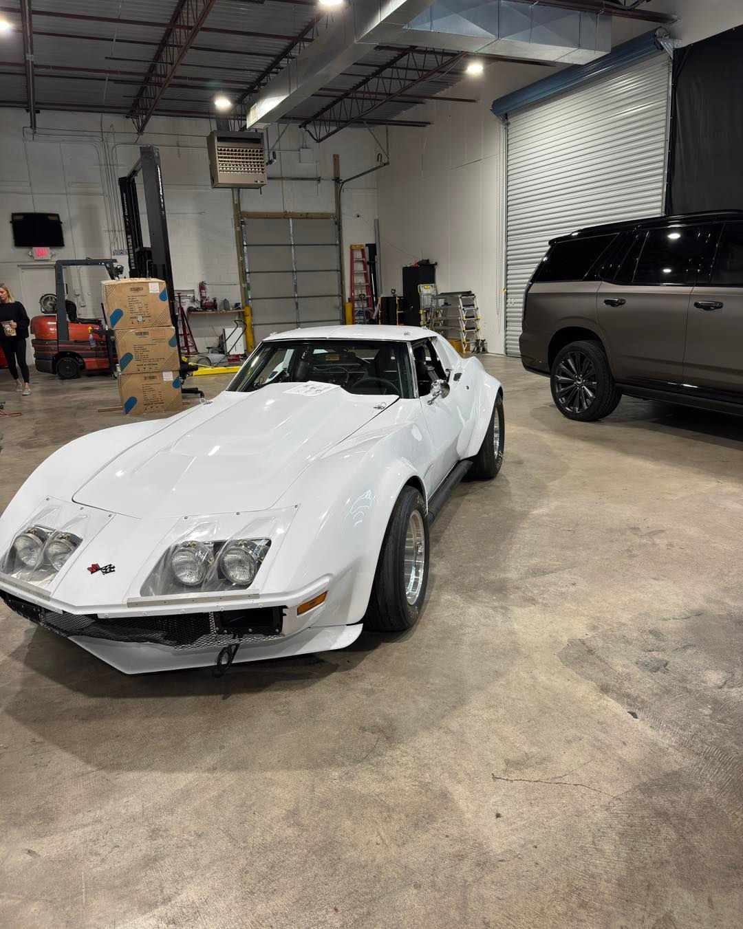 White sports car in a garage beside a gray SUV, with an open roll-up door in the background.