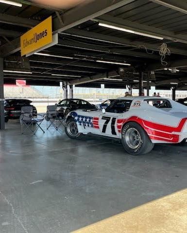 Vintage race car #76 in a garage or pit area, with other cars and a yellow sign overhead