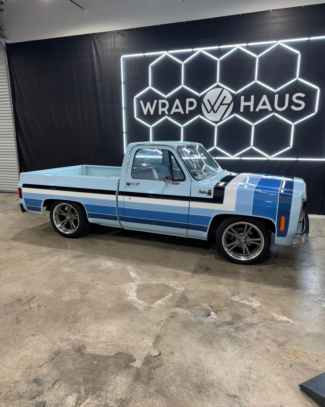 Blue vintage pickup truck parked in a shop with a glowing WRAP HAUS sign behind it