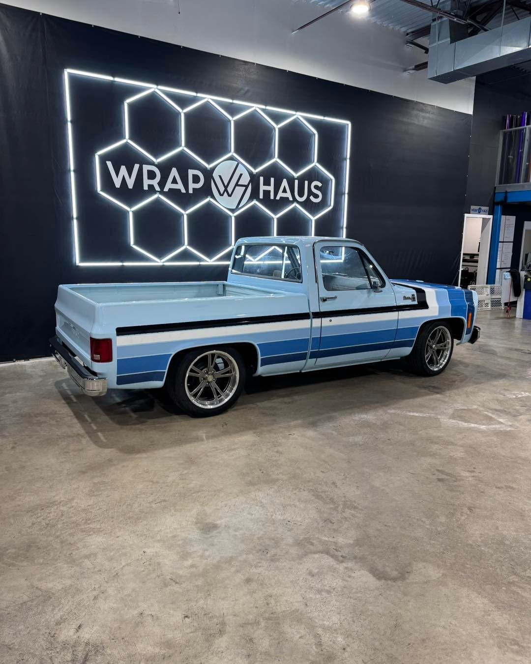 Custom light-blue pickup truck in a wrap shop with a glowing hexagon wall sign.