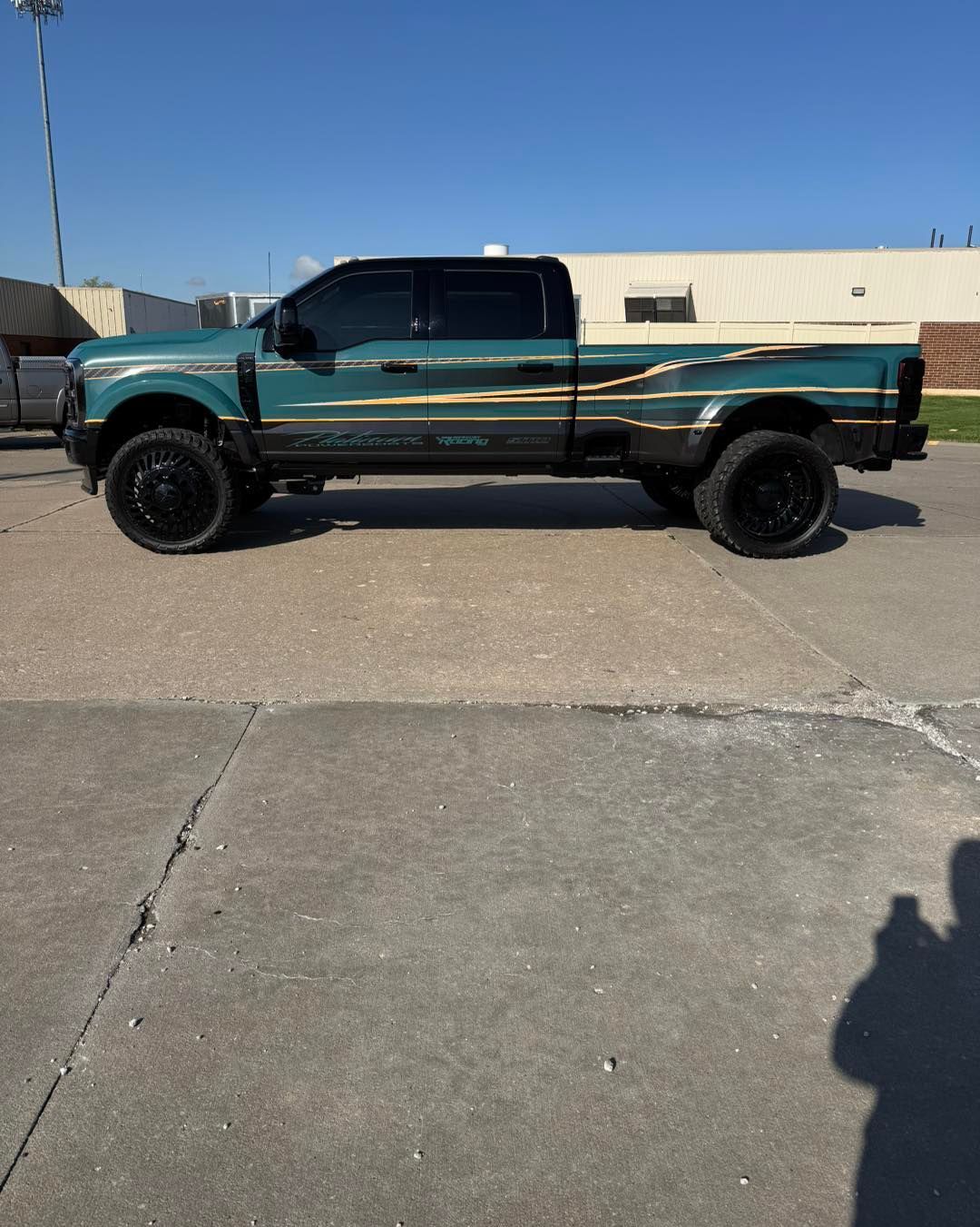 Teal and black lifted pickup truck parked on a street under a clear blue sky.
