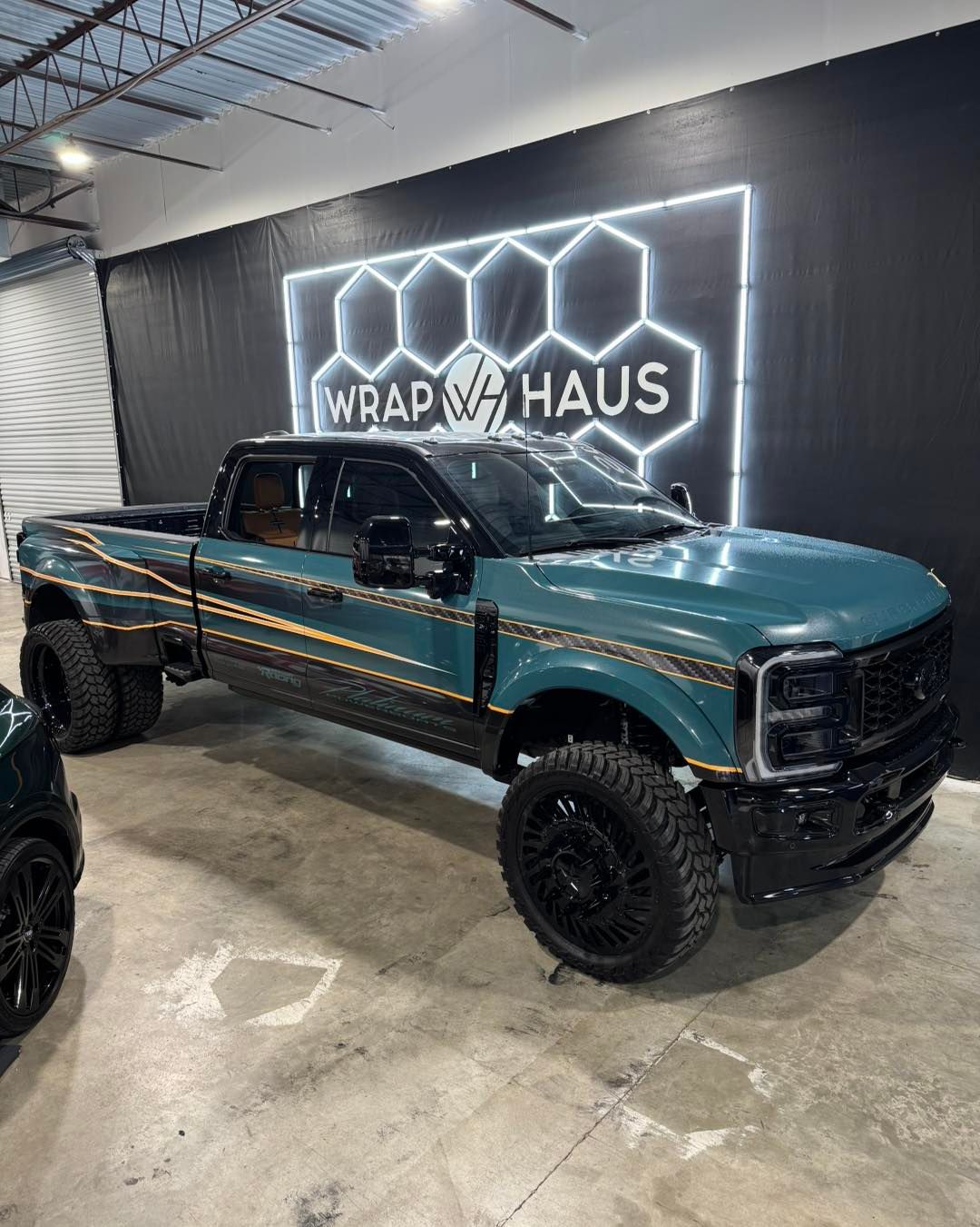 Teal lifted pickup truck with black accents in a garage, parked before a glowing WRAP HAUS sign