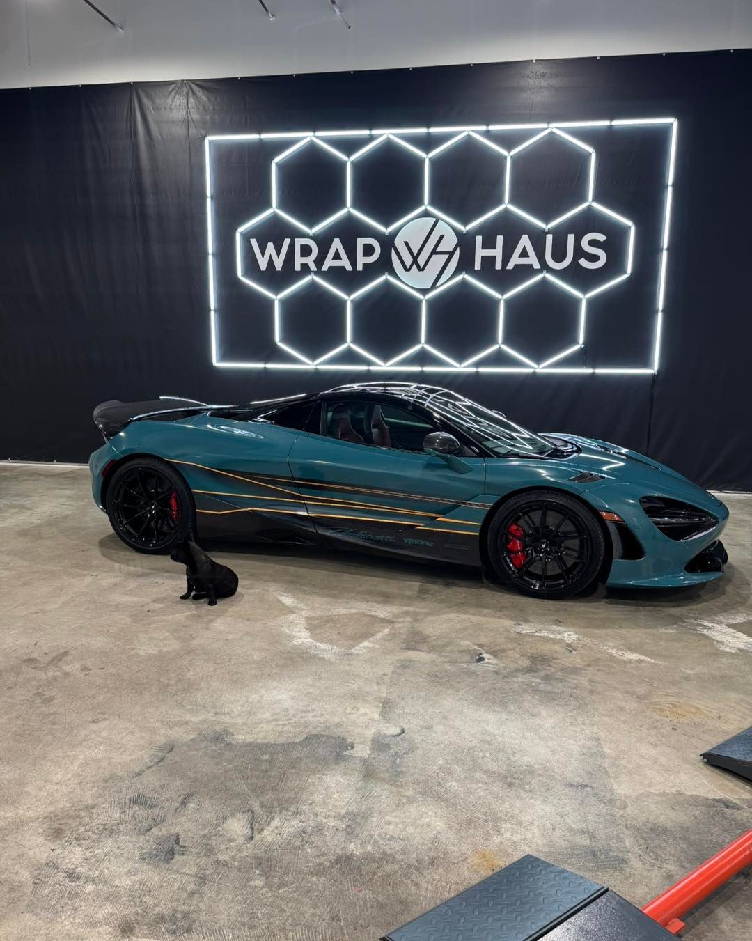 Teal sports car in a wrap shop, parked before a neon “WRAP HAUS” sign and hexagon wall display