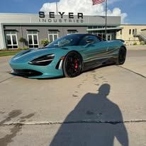 Teal McLaren sports car parked outside SEYER Industries on a sunny day, with a shadow in the foreground.