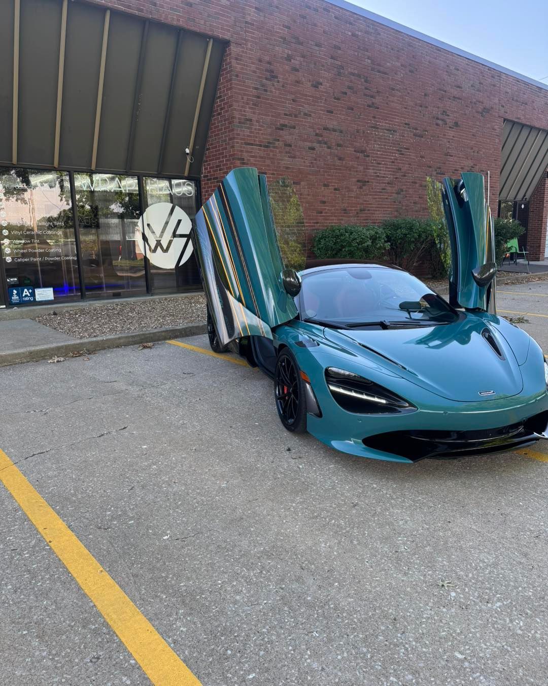 Teal sports car with butterfly doors open parked outside a brick building.