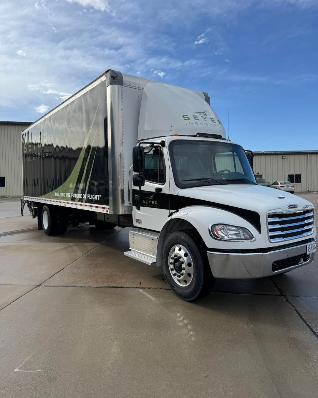 White delivery box truck parked on wet pavement outside a warehouse under a partly cloudy sky