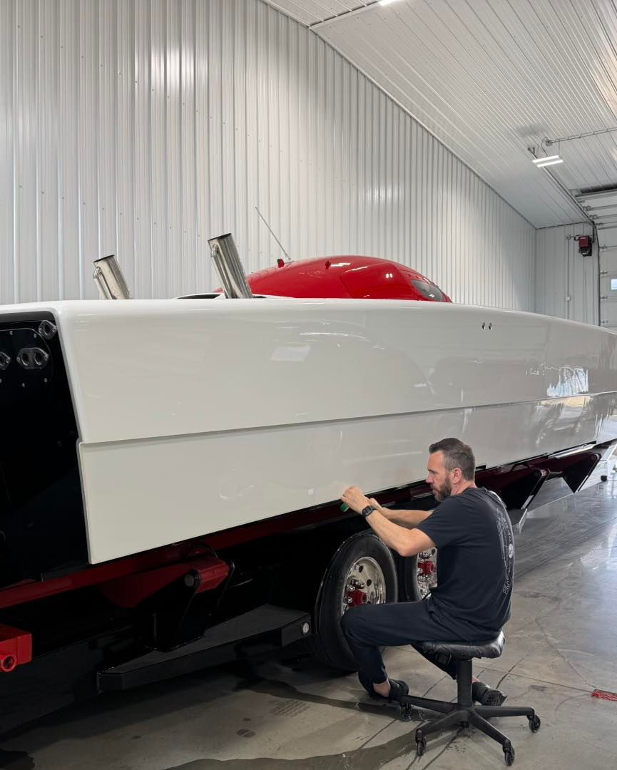 Man polishing the hull of a white speedboat in a workshop, with a red cover on top.