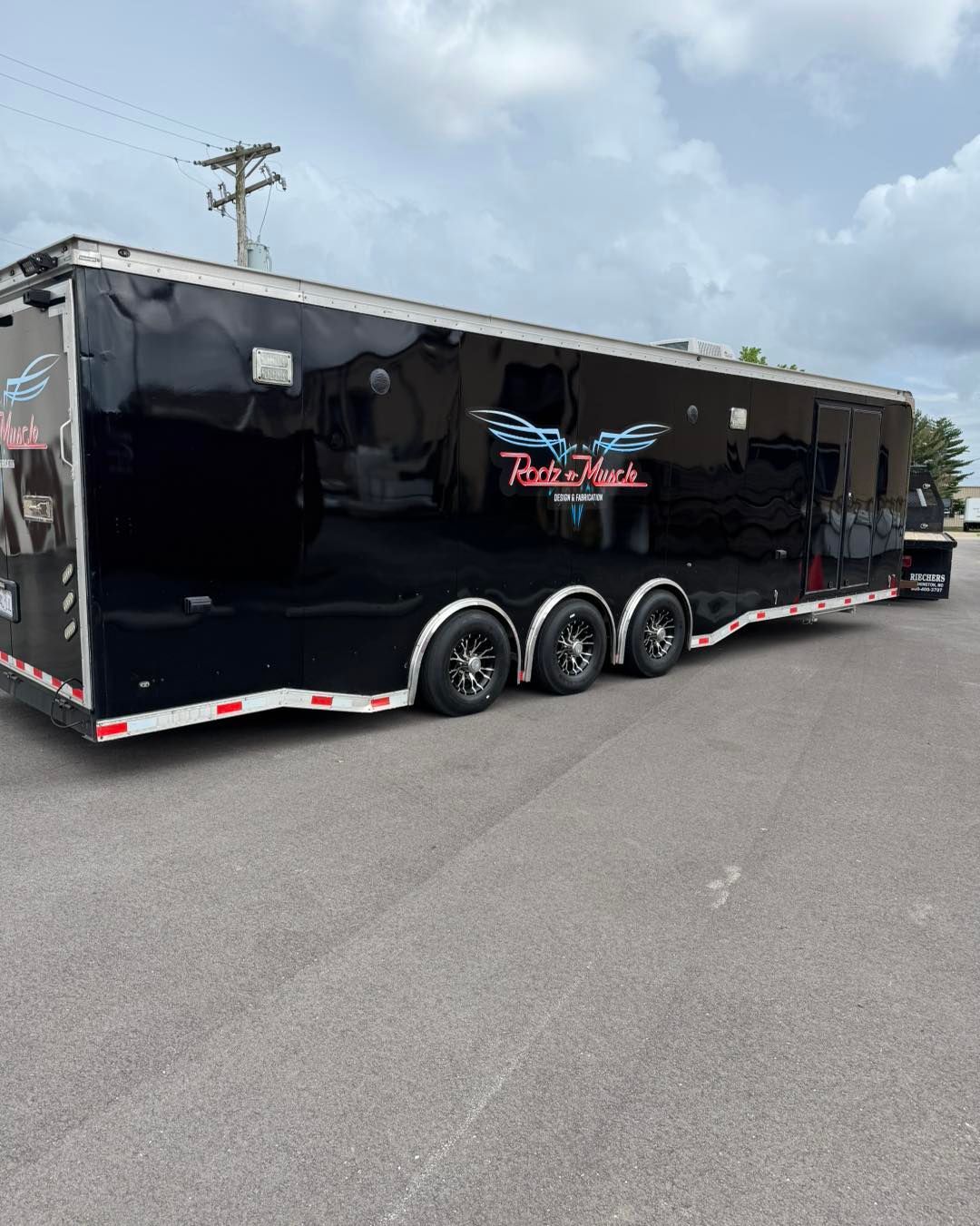 Black enclosed car trailer with three axles parked on pavement under a cloudy sky
