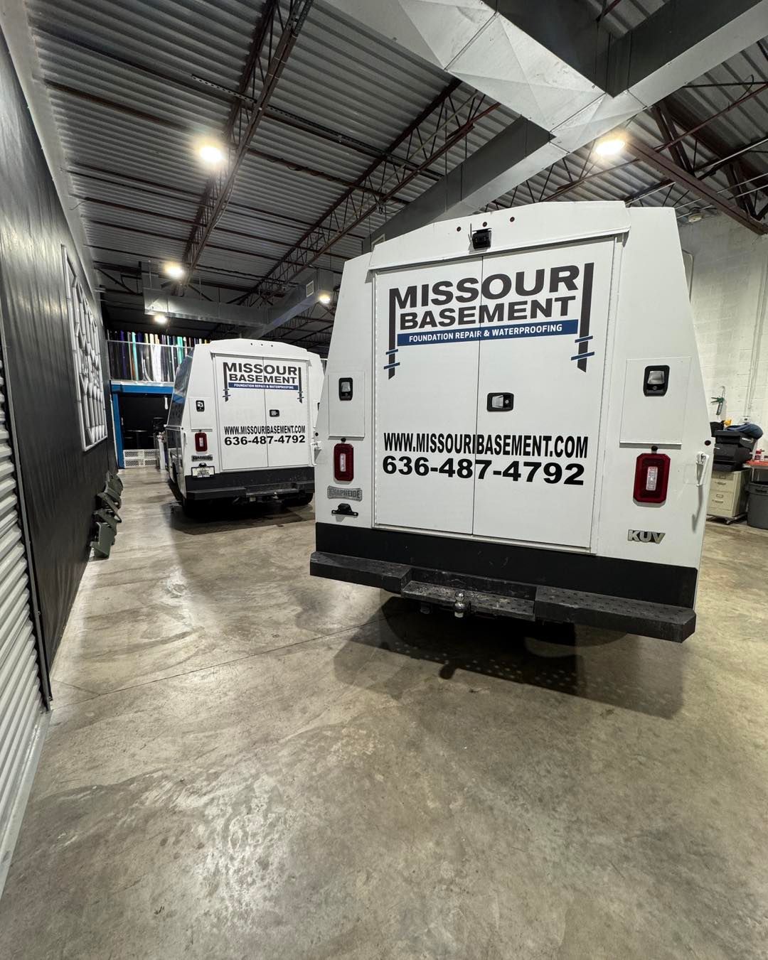 White Missouri Basement service vans parked in an indoor warehouse bay under bright lights.