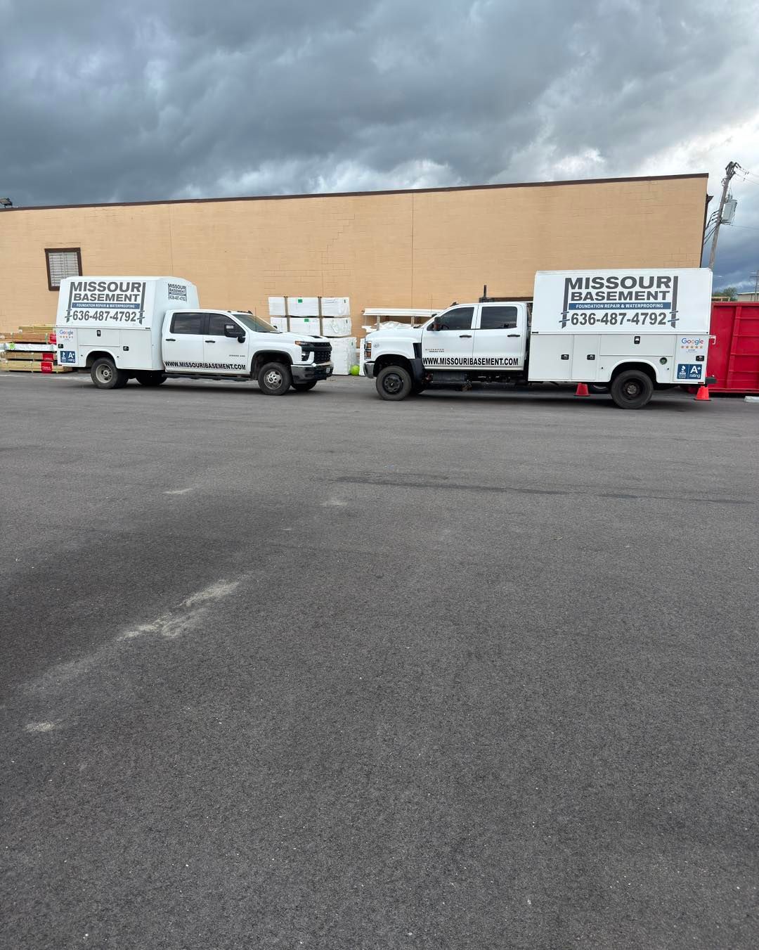 Two white utility trucks parked in front of a beige building under cloudy skies.