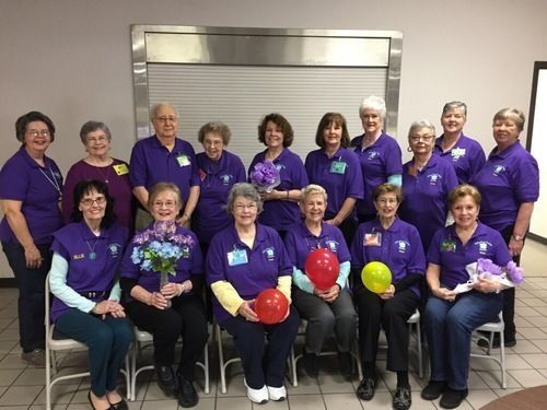 Group of people in purple shirts, holding balloons and flowers. They are in a room with a stainless steel surface.