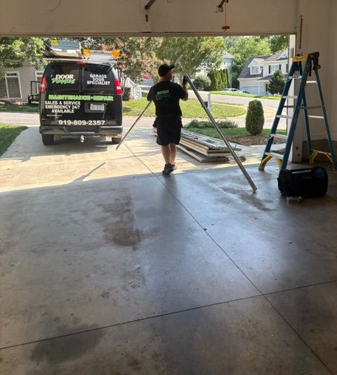 Worker carrying long poles into a garage from a driveway, with a work van outside.