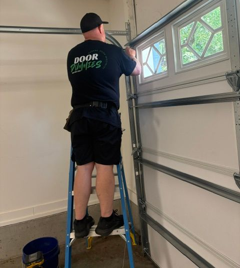 Worker installing or adjusting a garage door inside a garage, standing on a ladder.