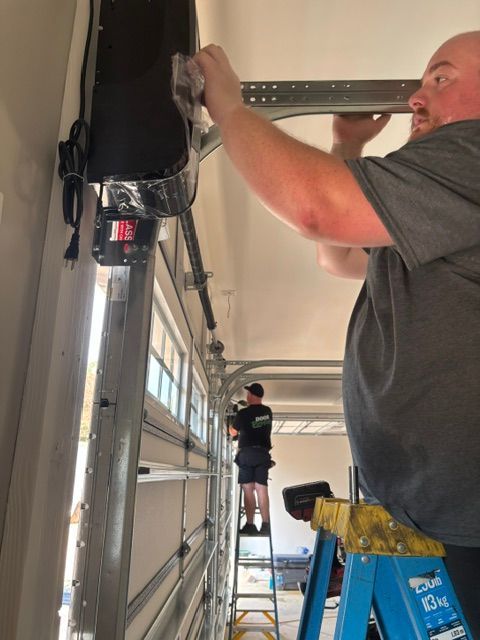 Two workers installing a garage door track, one on a ladder and one below with tools.