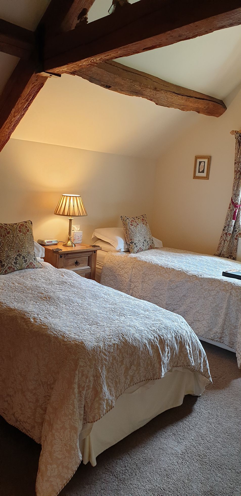 Bedroom with two twin beds, small bedside table, and decorative lamp. Room has sloped ceiling with exposed beams.