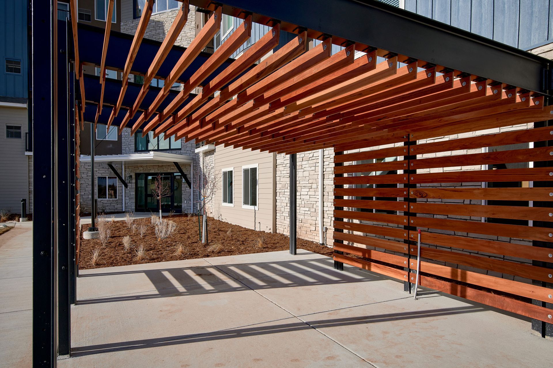 Covered walkway with orange slatted roof casting shadows onto a concrete courtyard beside a building
