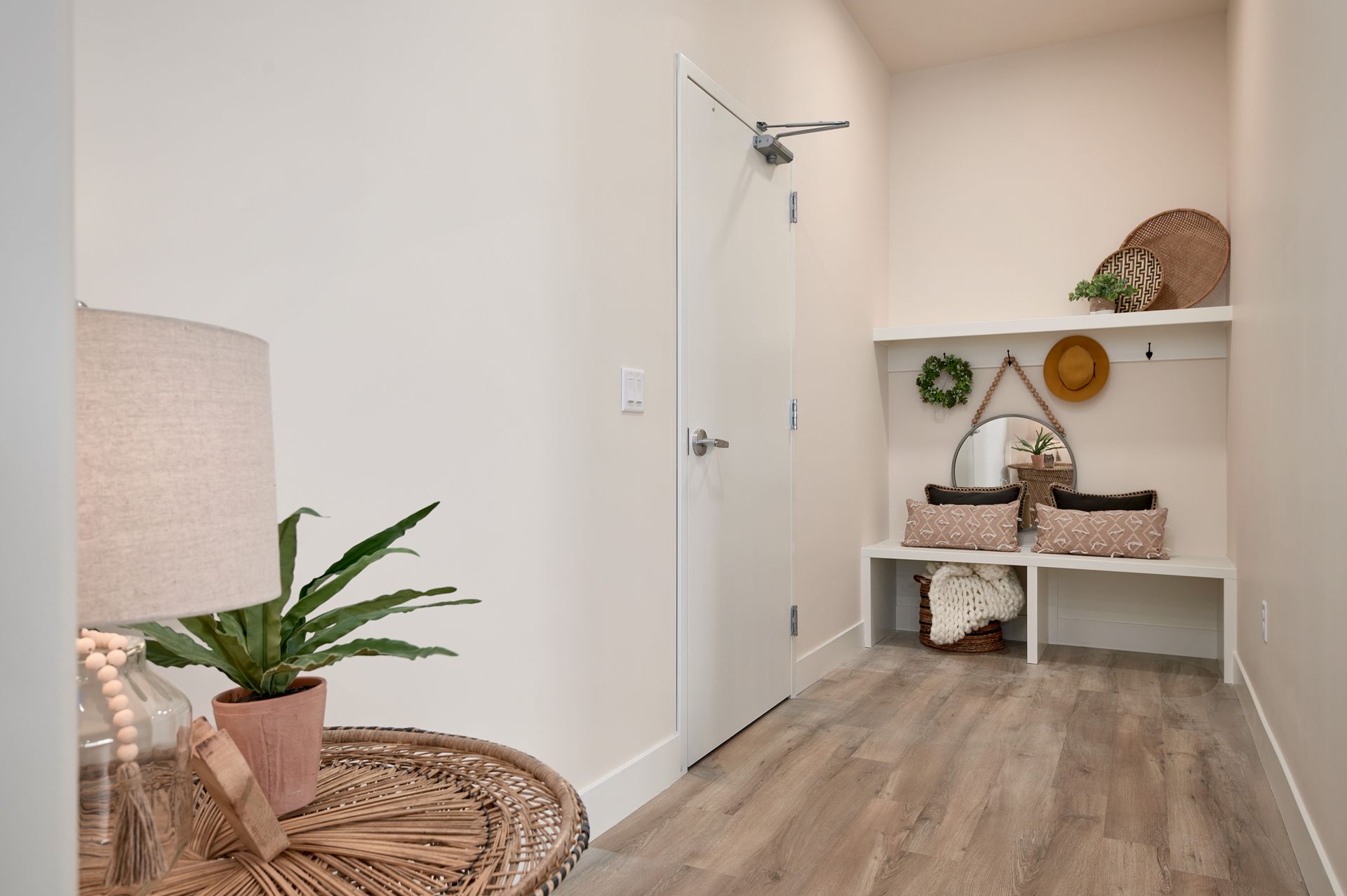 Small beige entryway with white walls, wood floor, bench, shelf decor, plant, and a woven table.
