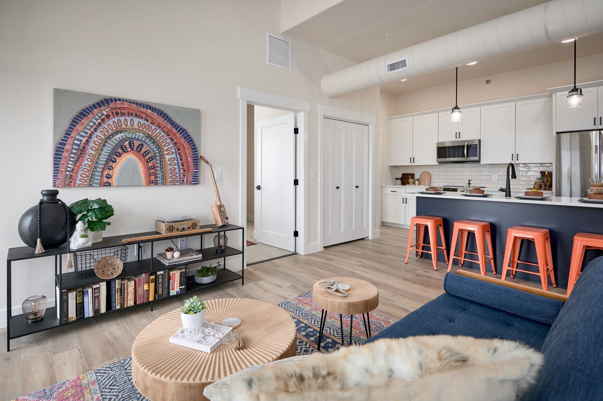 Bright modern living room and kitchen with blue sofa, orange stools, wall art, and a low console table