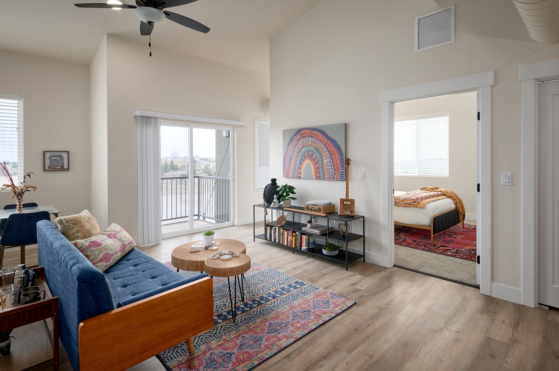 Bright living room with blue sofa, coffee table, colorful rug, TV stand, and open doorway to a bedroom