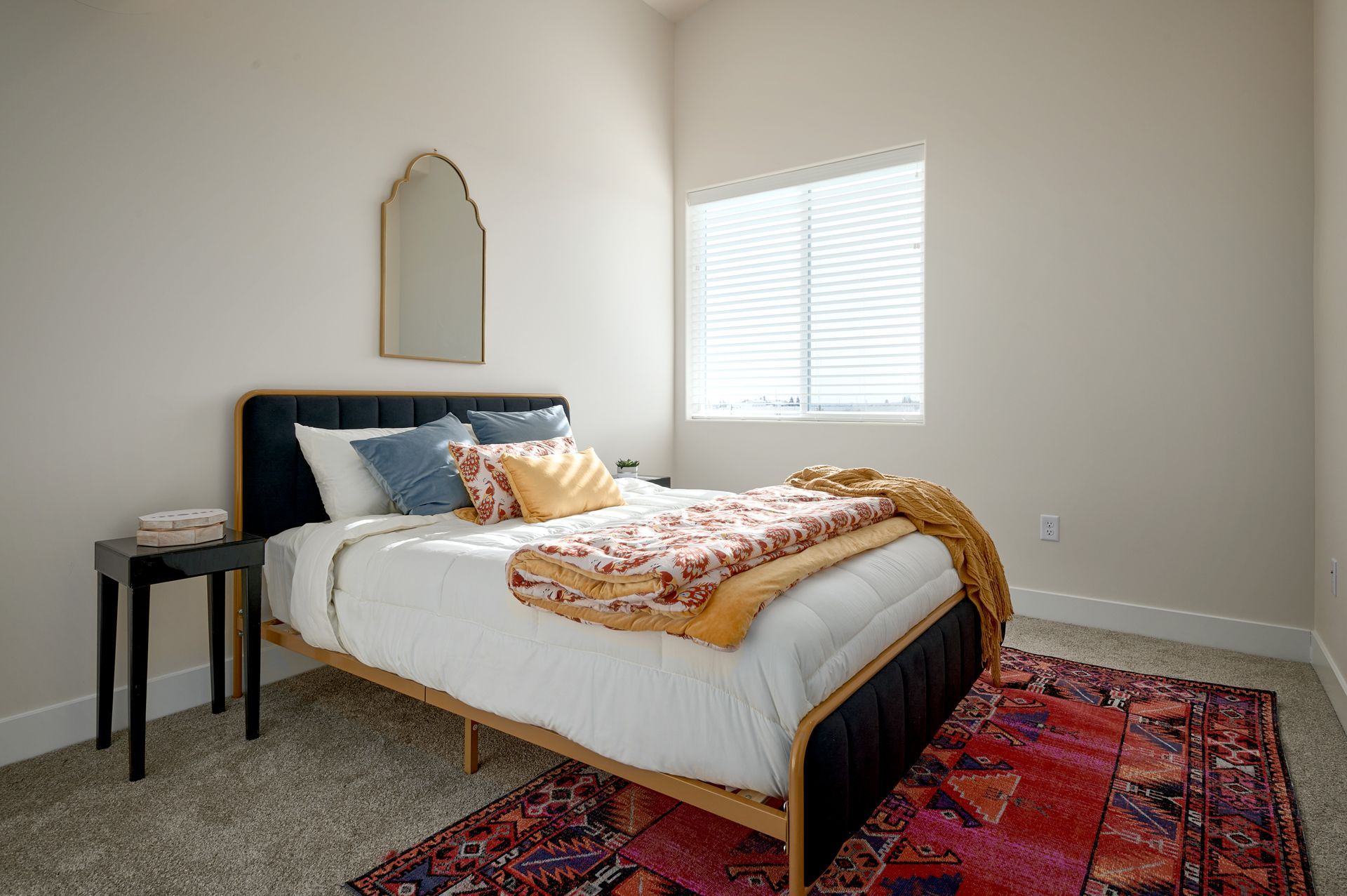 Bedroom with white bed, patterned rug, small side table, mirror, and window with blinds.
