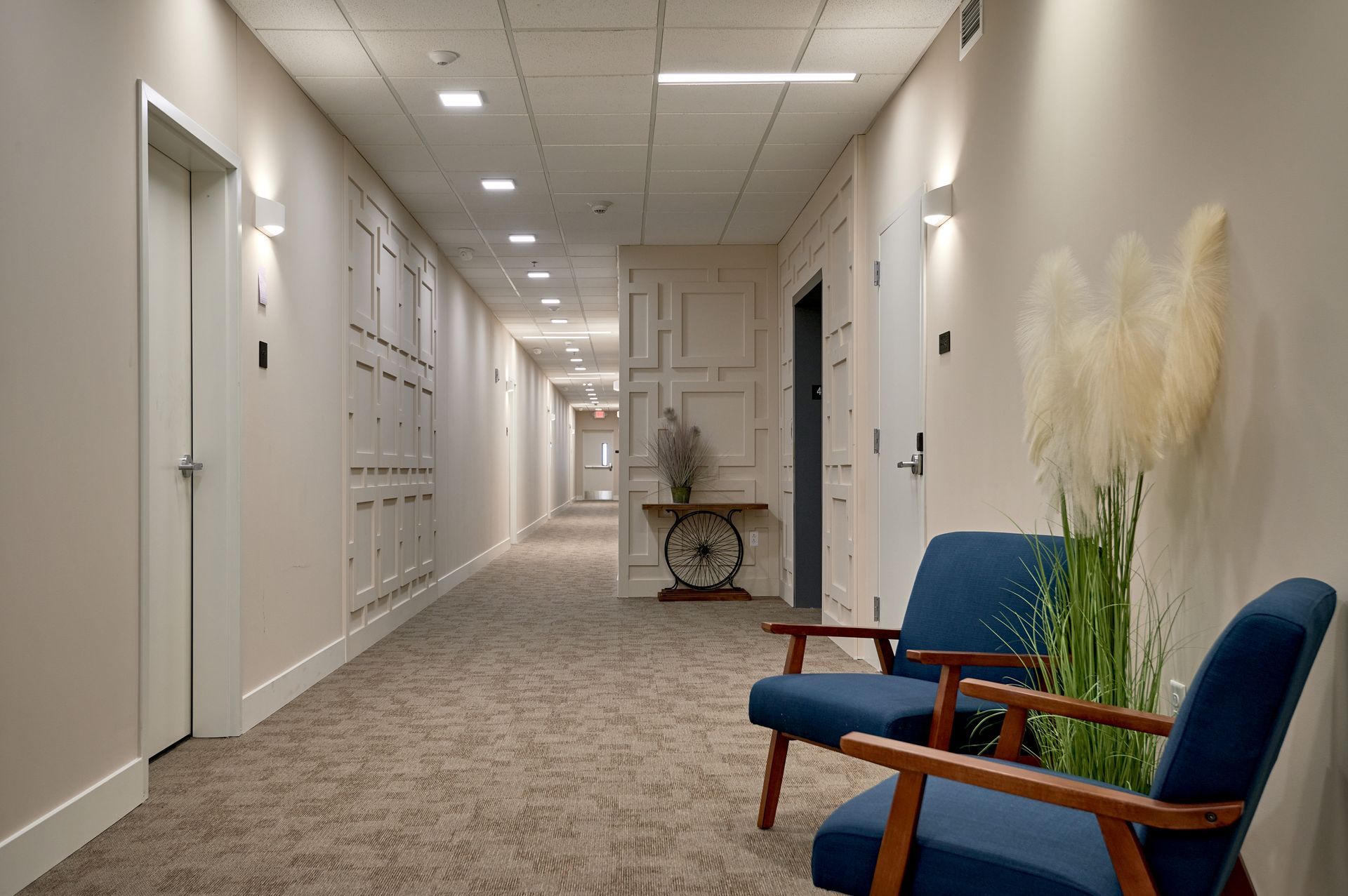 Quiet office hallway with lockers, blue chairs, and a plant on the right side.
