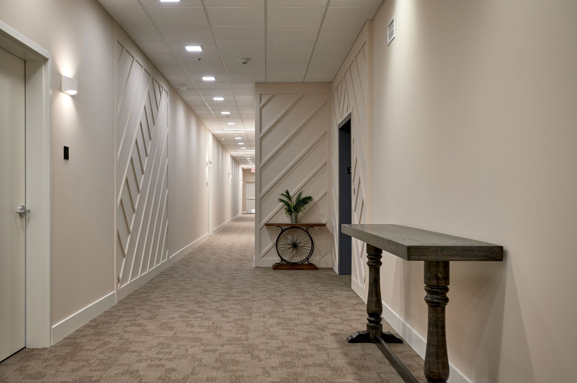 Empty beige hallway with carpet, wall sconces, decorative wall accents, and a small plant table