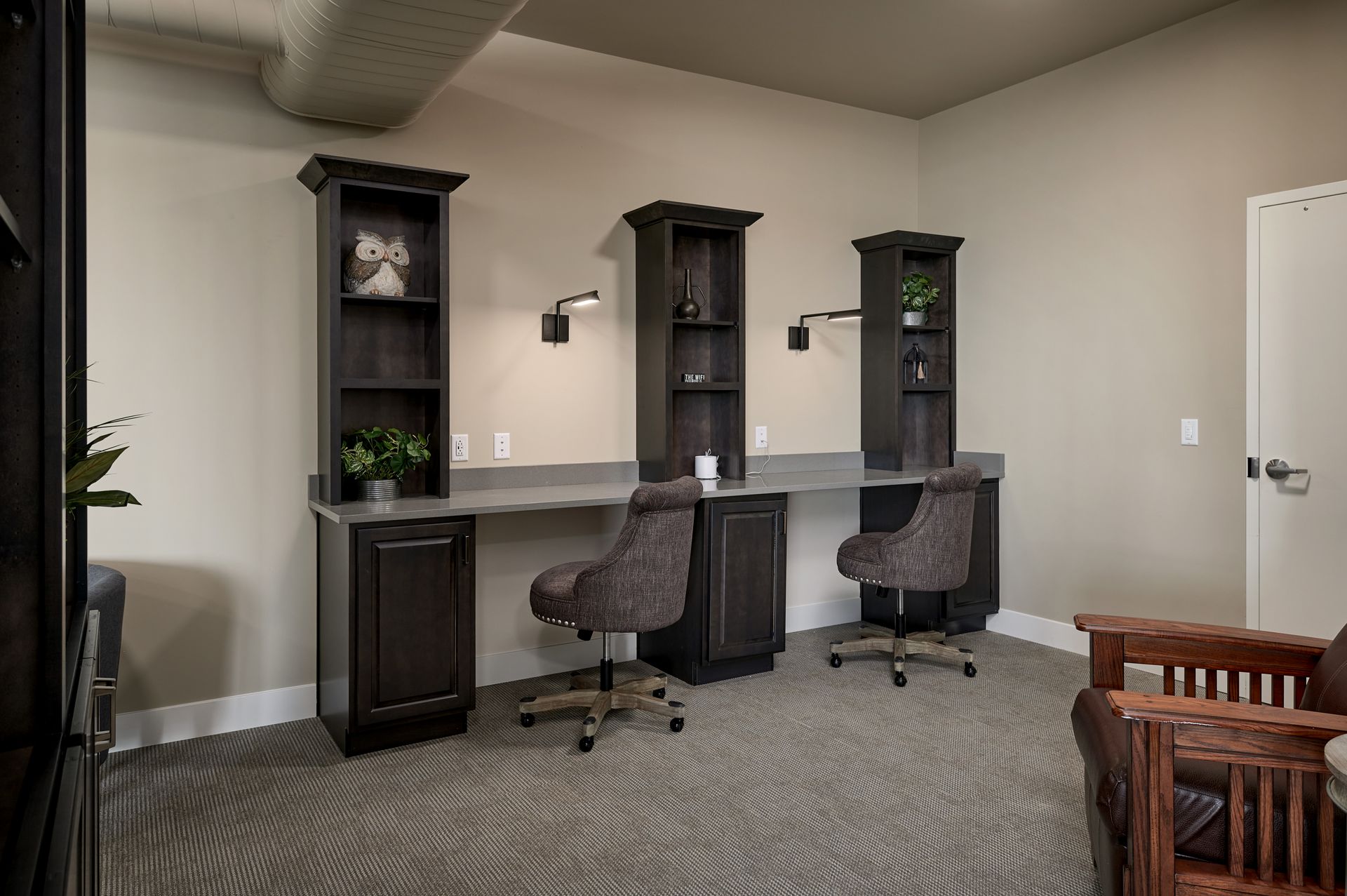 Modern home office with two desks, chairs, and dark wood shelving against beige walls