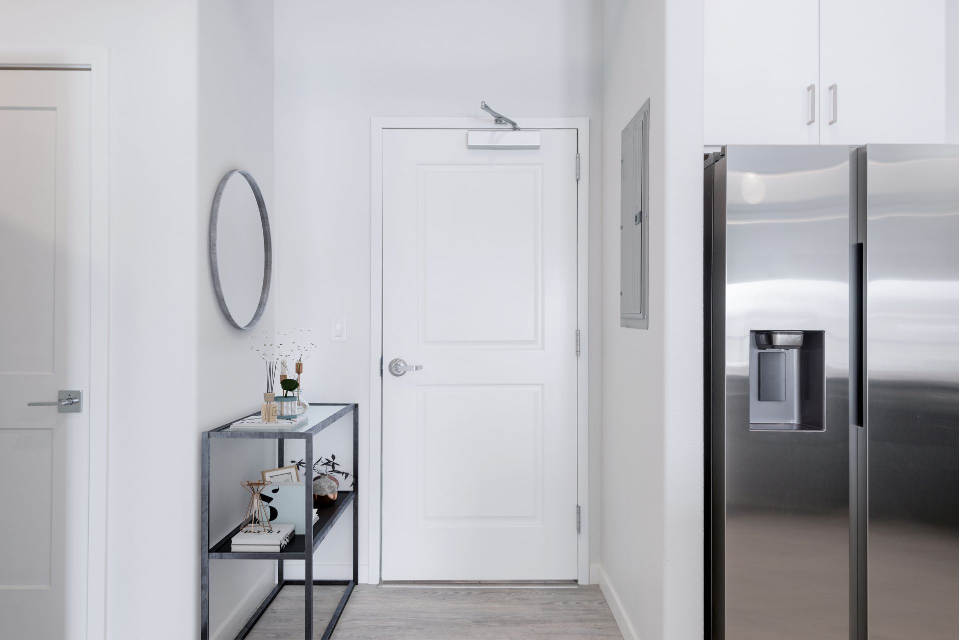 Bright white kitchen with stainless steel refrigerator, centered door, and small wall shelf with mirror.