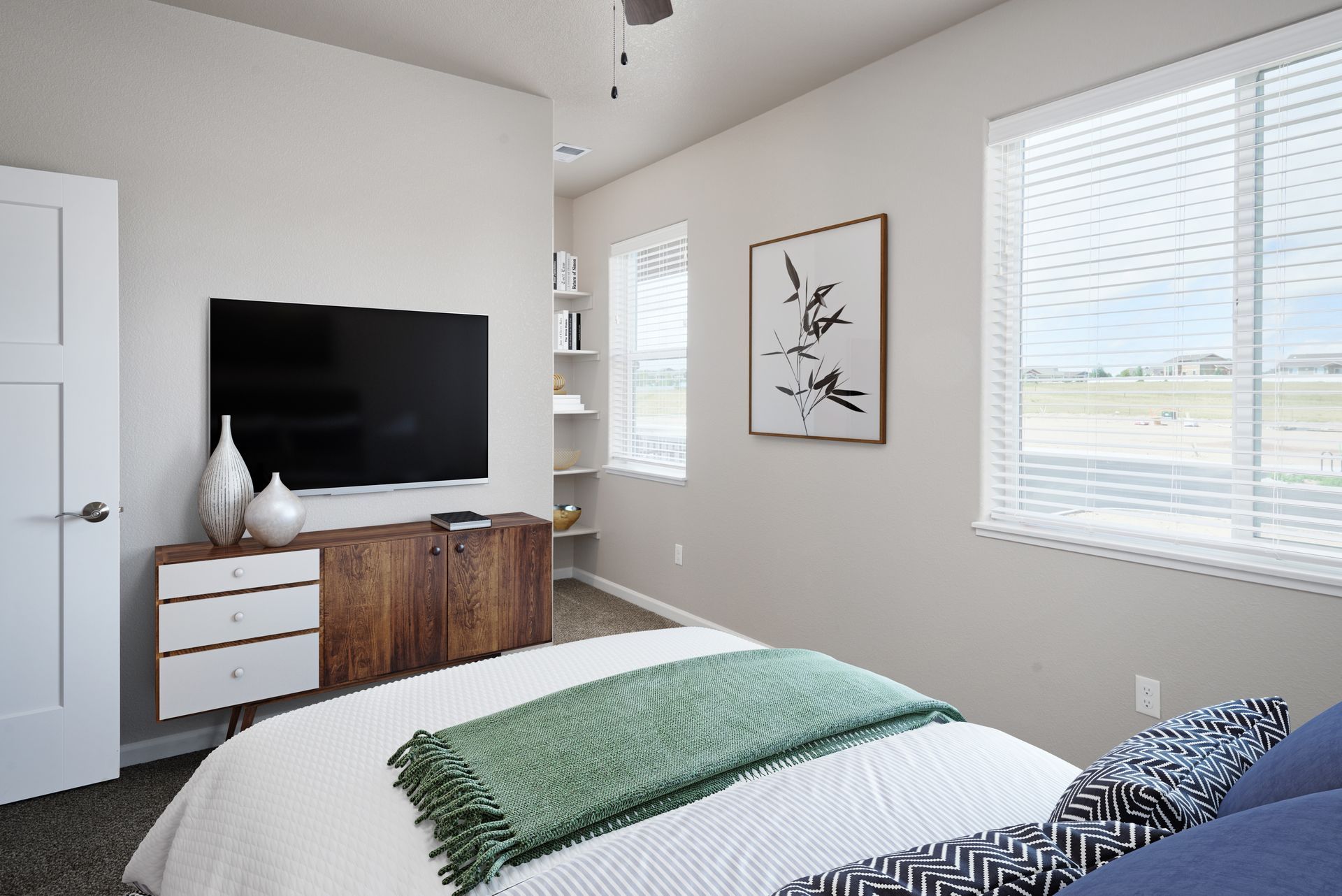Bright bedroom with TV, wood dresser, white blinds, wall art, and a bed with a green blanket.