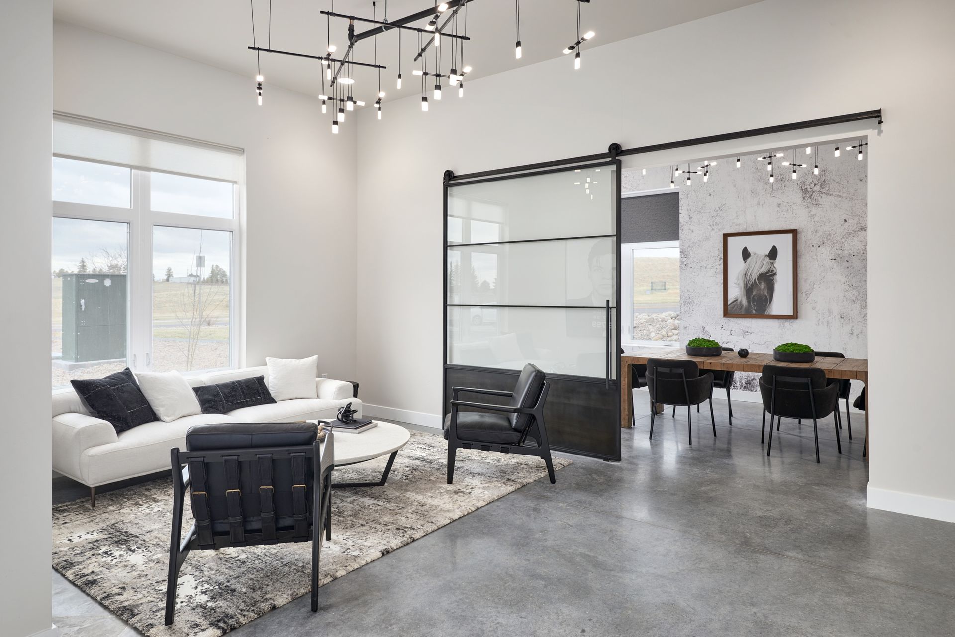 Modern open-plan living room and dining area with white sofa, black chairs, glass partition, and gray concrete floor